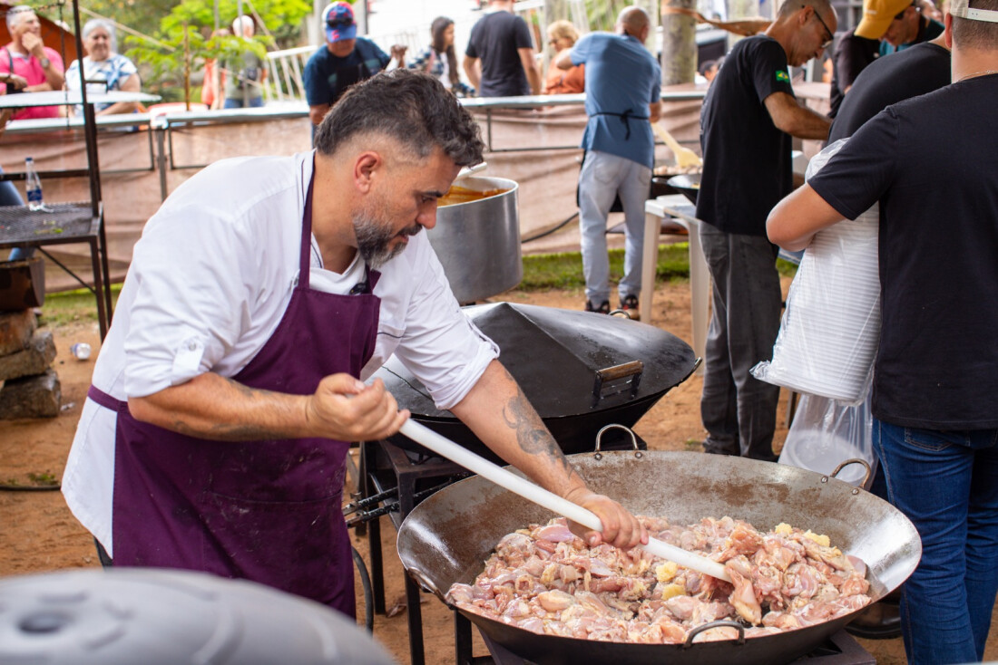 Um cozinheiro de avental roxo mexe grandes pedaços de frango em um tacho de metal gigante durante evento ao ar livre em Guararema. Ao fundo, outras pessoas trabalham no preparo e o público aguarda em uma área festiva.