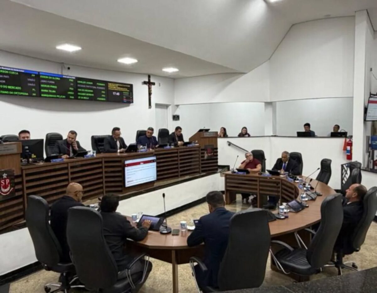 Vista ampla do plenário da Câmara Municipal de Santa Isabel durante uma sessão. Vereadores estão sentados em bancadas de madeira semicirculares com computadores e tablets, debatendo e acompanhando a votação. Ao fundo, um grande painel digital exibe informações com luzes verdes e uma cruz de madeira está fixada na parede branca.