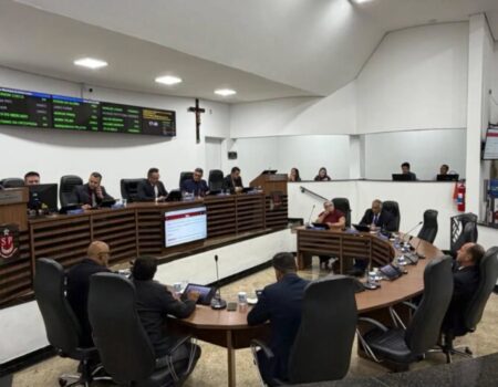 Vista ampla do plenário da Câmara Municipal de Santa Isabel durante uma sessão. Vereadores estão sentados em bancadas de madeira semicirculares com computadores e tablets, debatendo e acompanhando a votação. Ao fundo, um grande painel digital exibe informações com luzes verdes e uma cruz de madeira está fixada na parede branca.