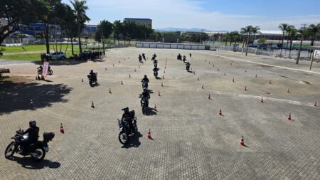 Vista aérea de um amplo pátio de paralelepípedos em Poá, onde diversos agentes da GCM em motocicletas pretas e uniformizados realizam manobras práticas em um percurso delimitado por cones laranjas e brancos, sob um céu claro. Ao fundo, árvores e prédios da cidade.